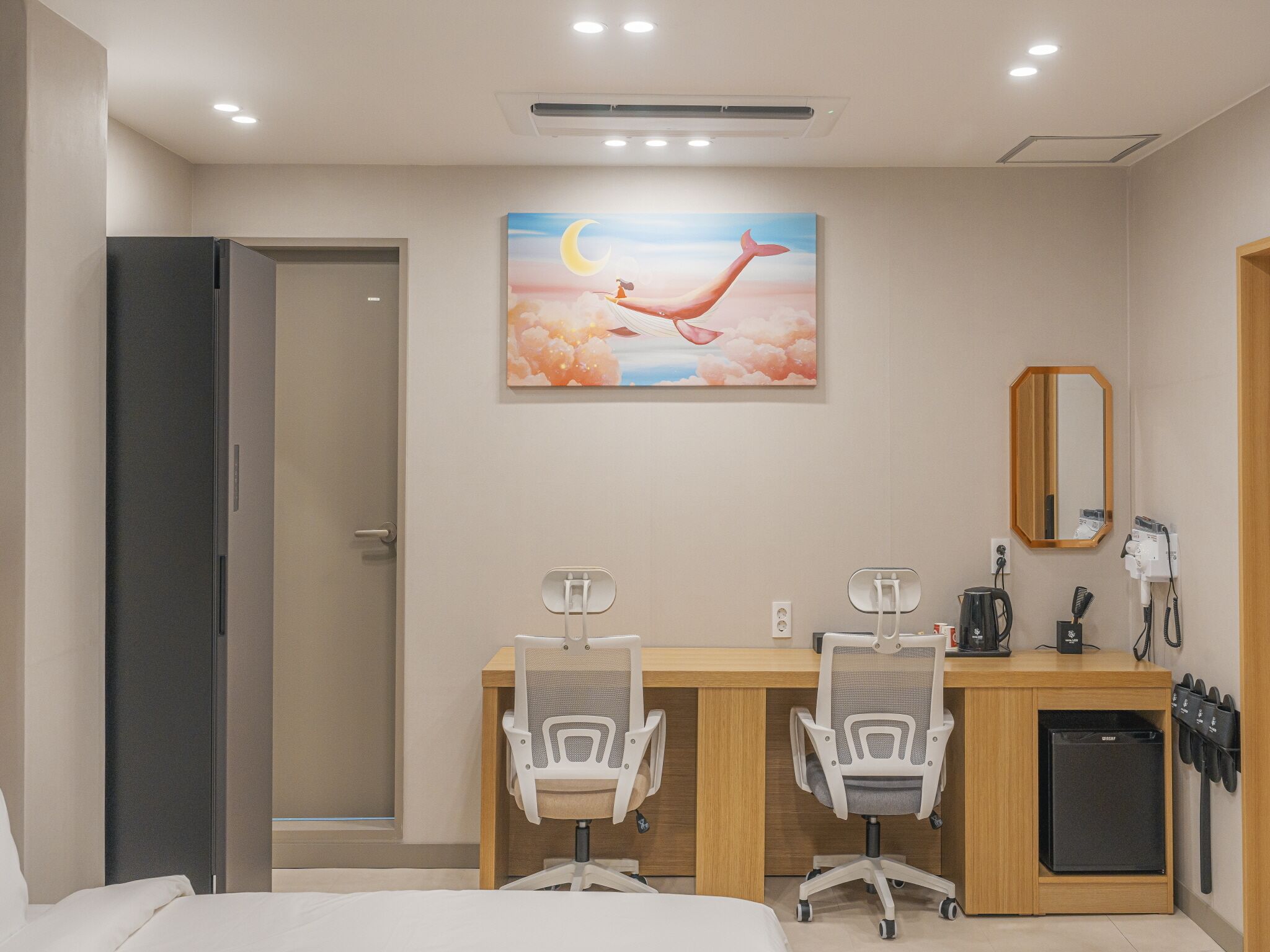 A hotel room with a bed, a desk with two chairs, a small refrigerator, a hairdryer, and a painting of a bird on the wall.