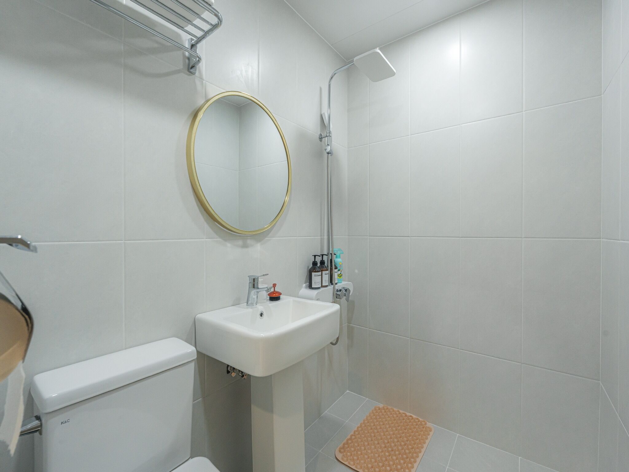 Une salle de bain comprenant des toilettes, un lavabo, un miroir, une douche et un petit tapis sur le sol.