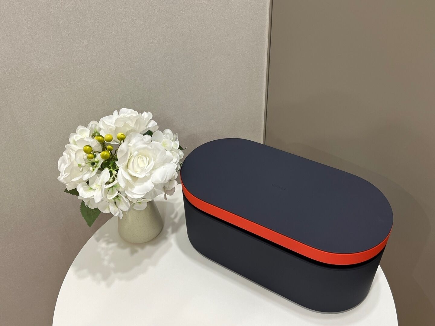 A white vase with white flowers and a black and red box on a white surface.