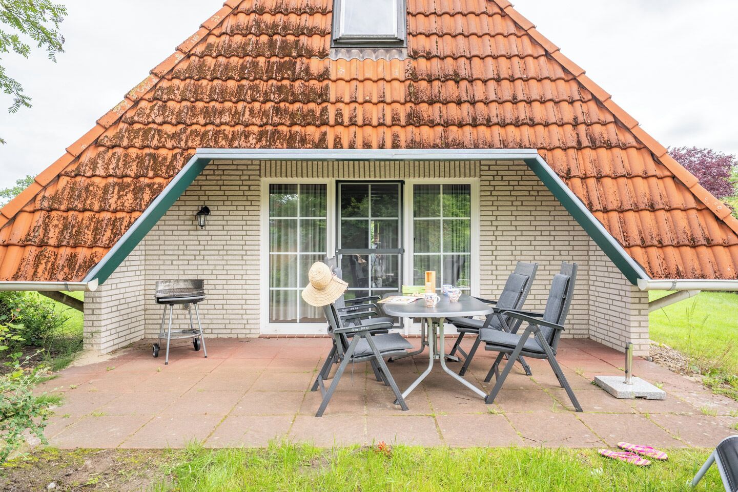 A house with a sloped roof, a patio with a table and chairs, and a grill.