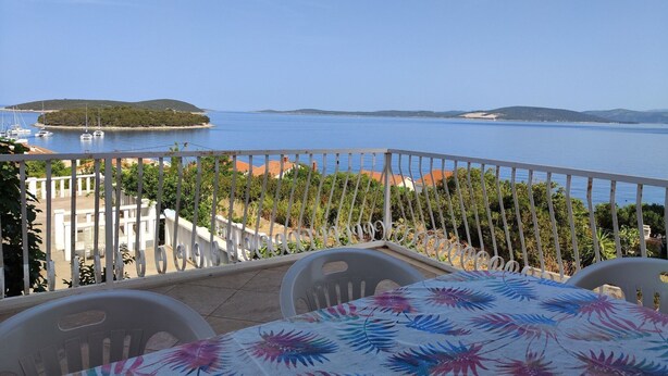Un balcon avec une table et des chaises, donnant sur une mer calme où l’on peut apercevoir des bateaux et une île.