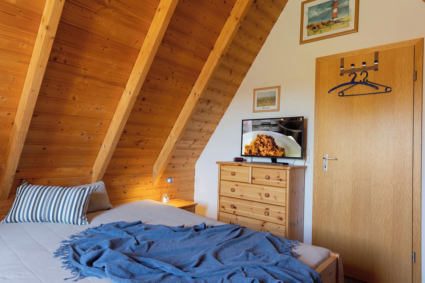 A bedroom with a sloped ceiling, wooden beams, a bed with a blue blanket, a dresser, a television on top, and a door with a hanger.