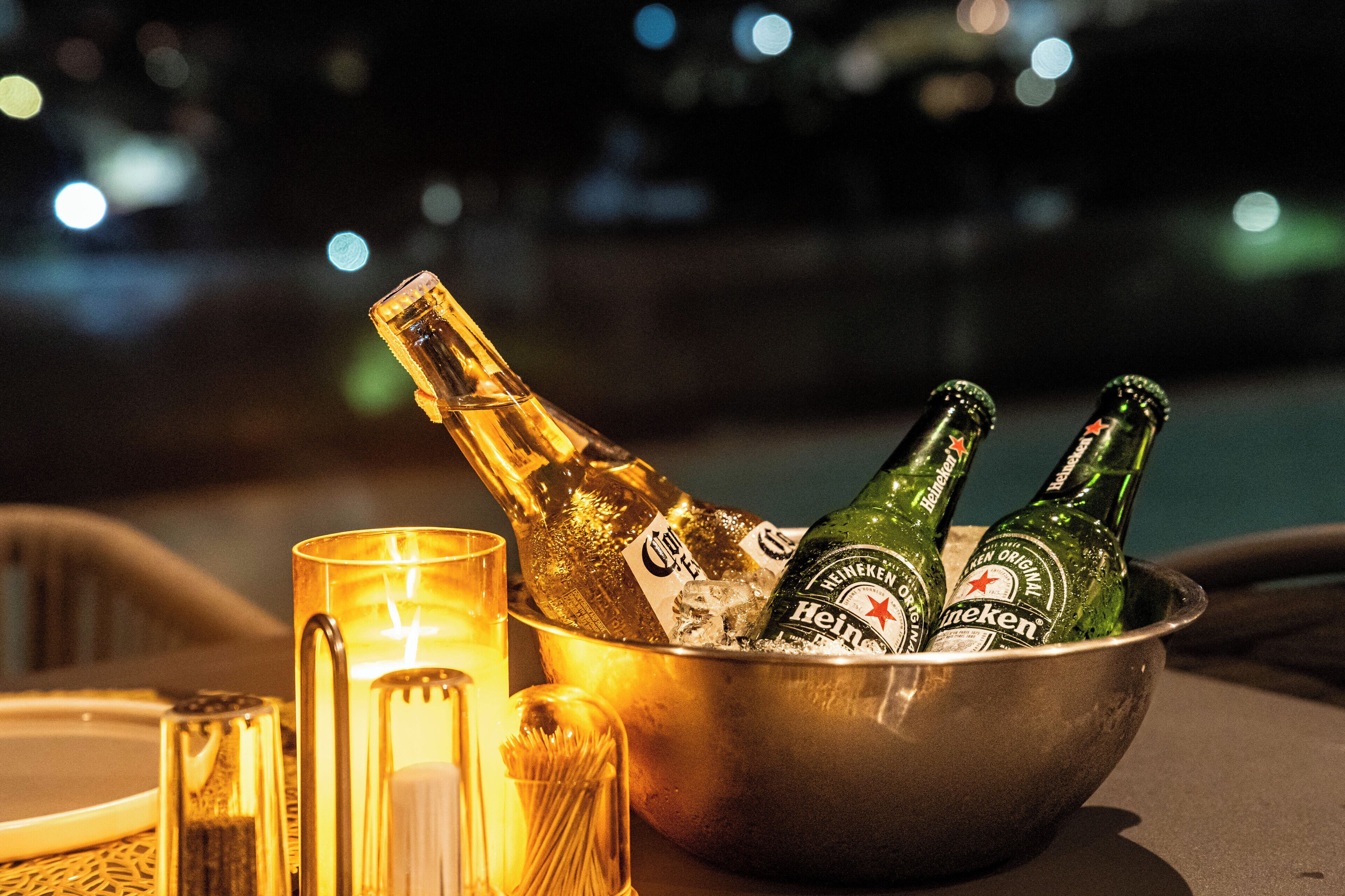 A metal bowl containing bottles of Heineken beer and a glass of champagne, with a candle and a glass on a table.