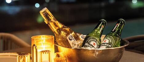 A metal bowl containing bottles of Heineken beer and a glass of champagne, with a candle and a glass on a table.