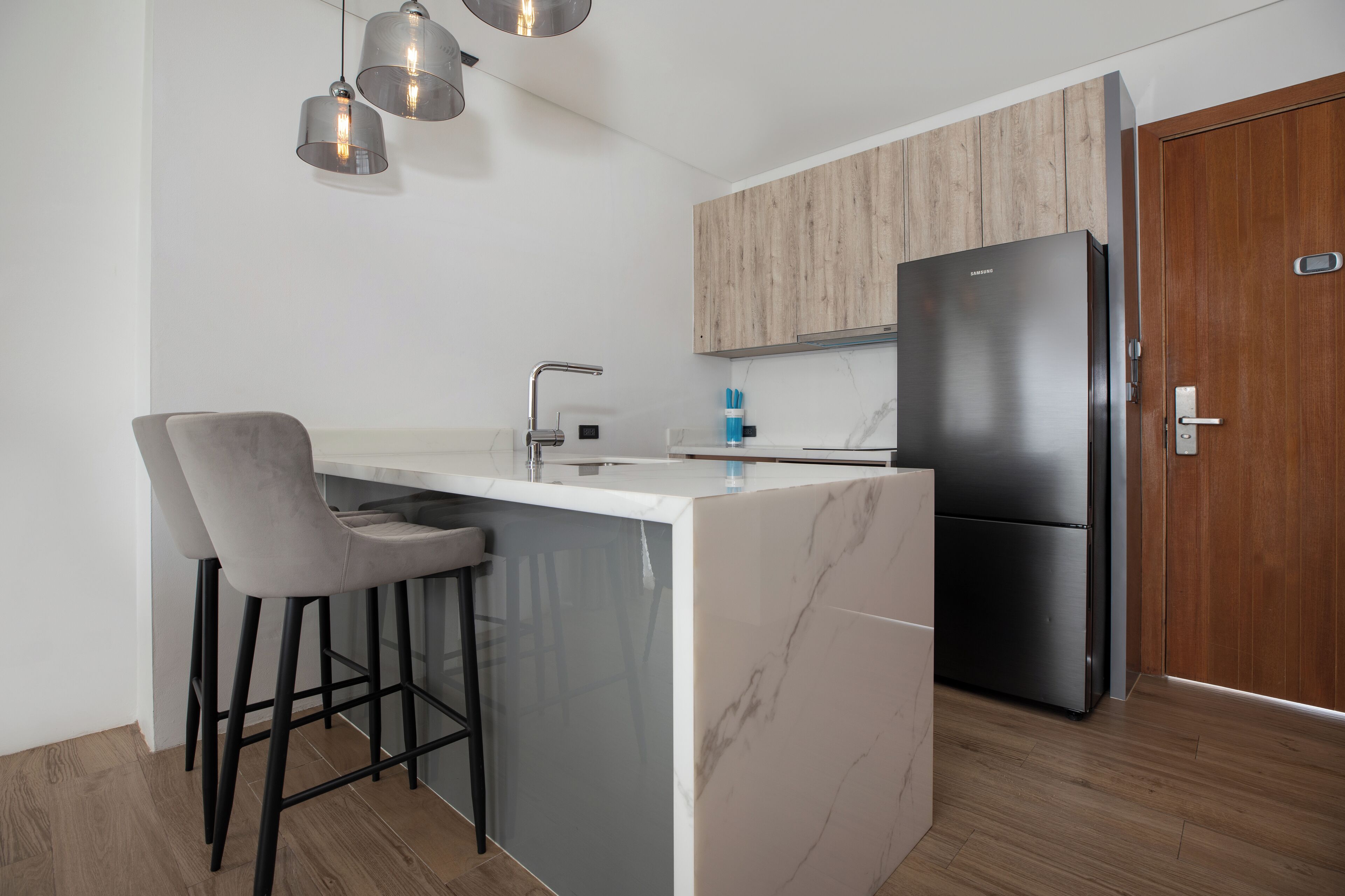 A modern kitchen with a marble island, two bar stools, a stainless steel refrigerator, and wooden cabinets.