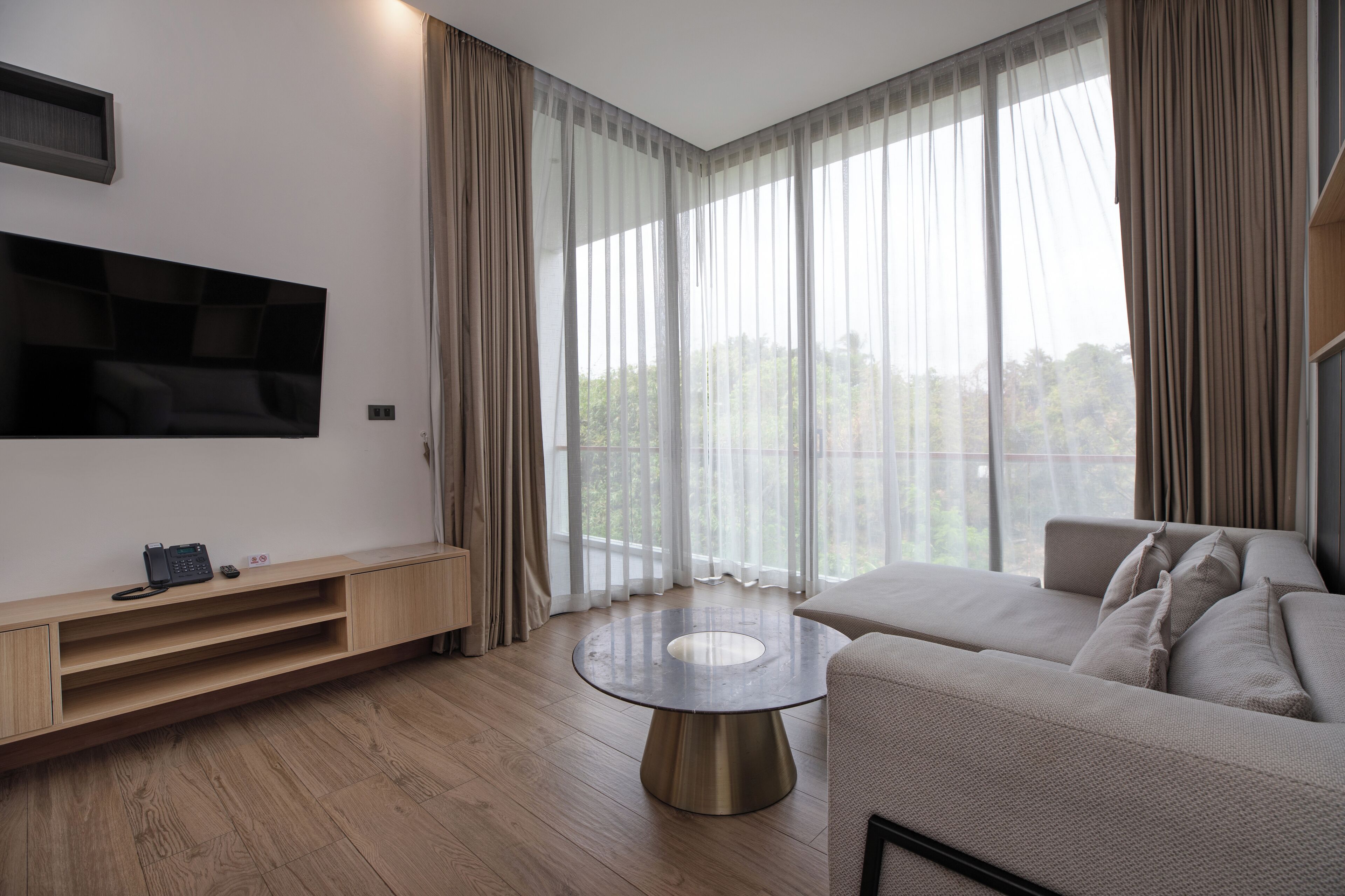 A modern living room with a flat-screen TV mounted on the wall, a round coffee table, a beige sofa, and a large window with sheer curtains.