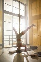 Una persona haciendo una postura de equilibrio sobre una esterilla de yoga en una habitación con ventanas grandes y suelo de madera.