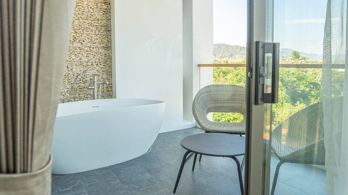 Ein modernes Badezimmer mit einer freistehenden Badewanne, einem Korbstuhl und einem Balkon mit Blick auf Grünflächen.