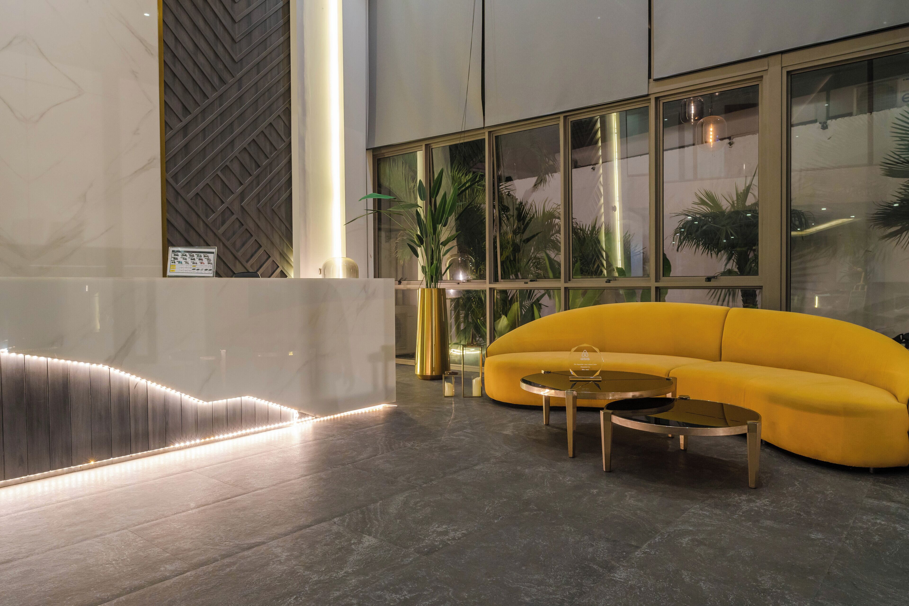 A modern lobby with a curved yellow sofa, two round coffee tables, and a large window with plants.