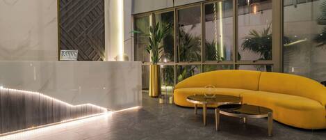 A modern lobby with a curved yellow sofa, two round coffee tables, and a large window with plants.
