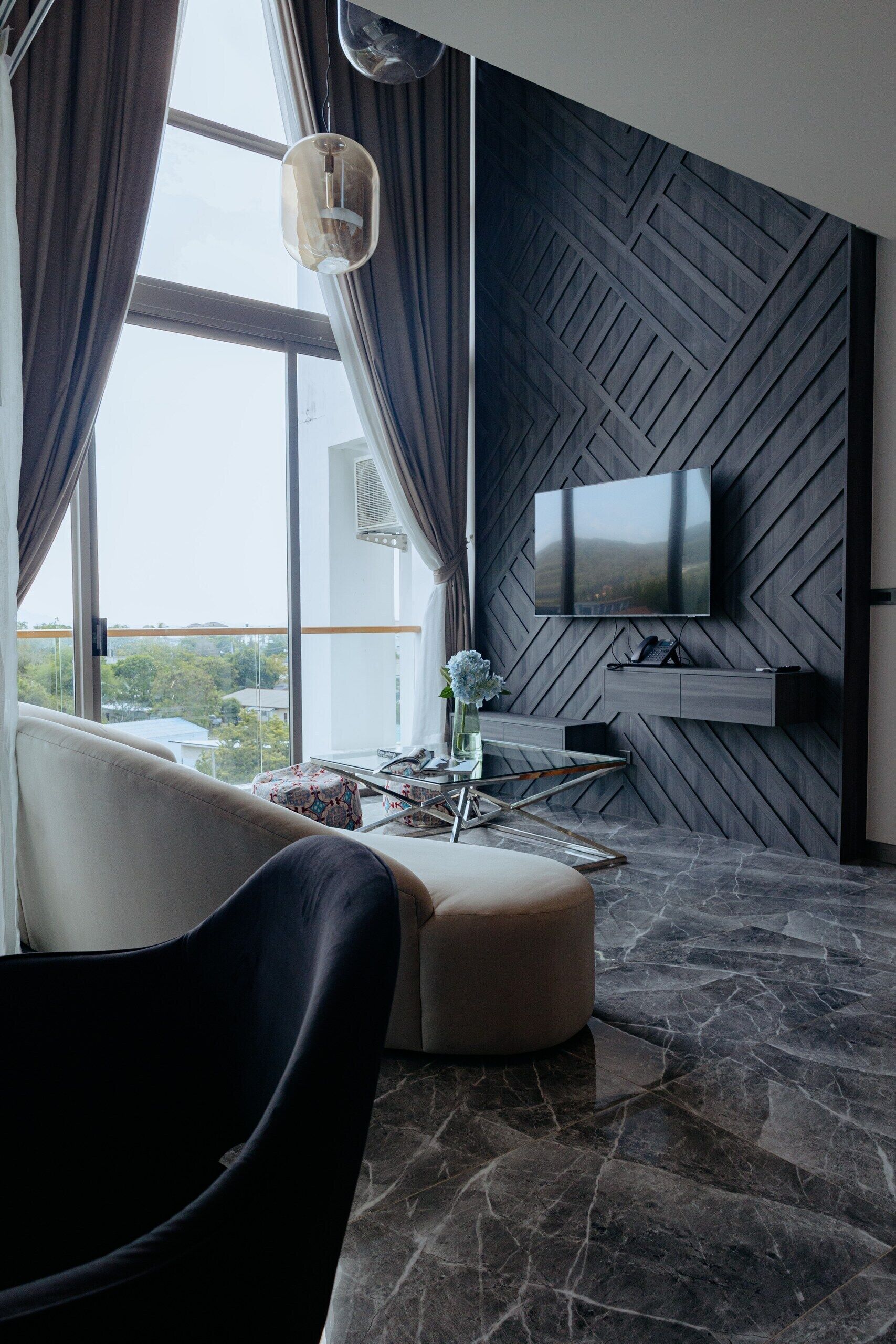 A modern living room with a large window, a flat-screen TV mounted on a geometric wall, a black chair, a beige ottoman, and a glass coffee table.