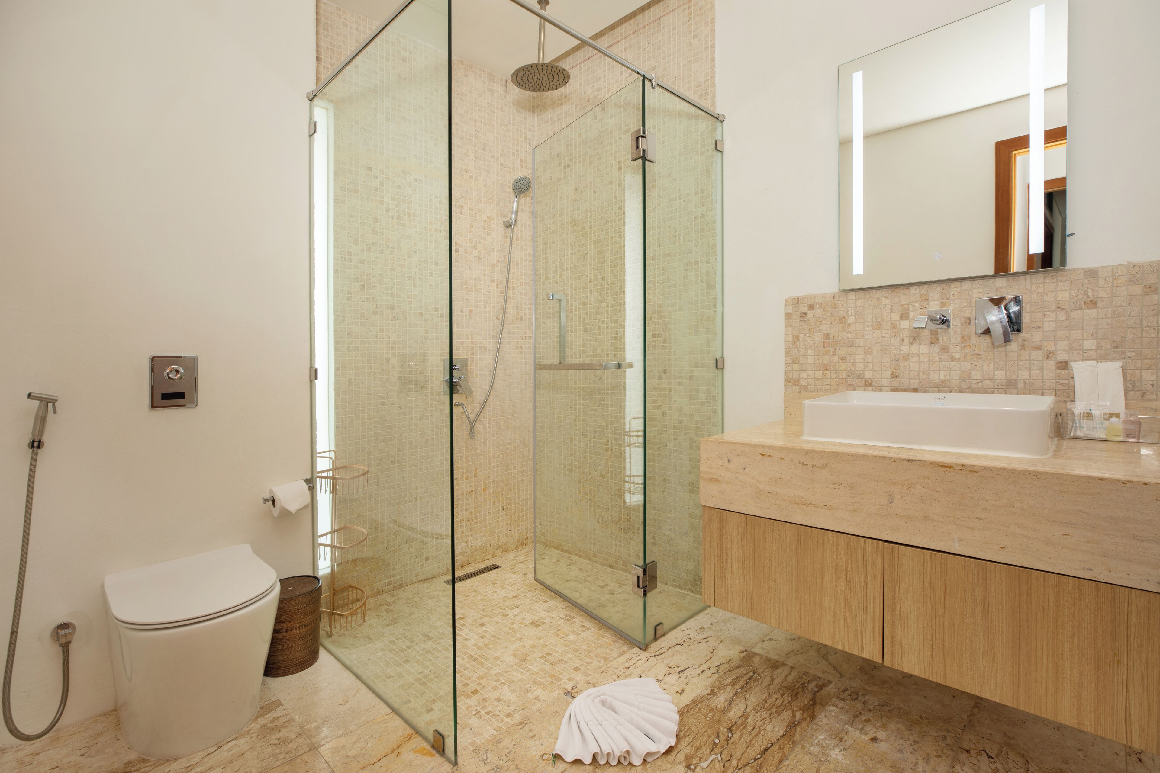 A modern bathroom with a glass-enclosed shower, a white toilet, and a beige-tiled vanity with a large mirror.