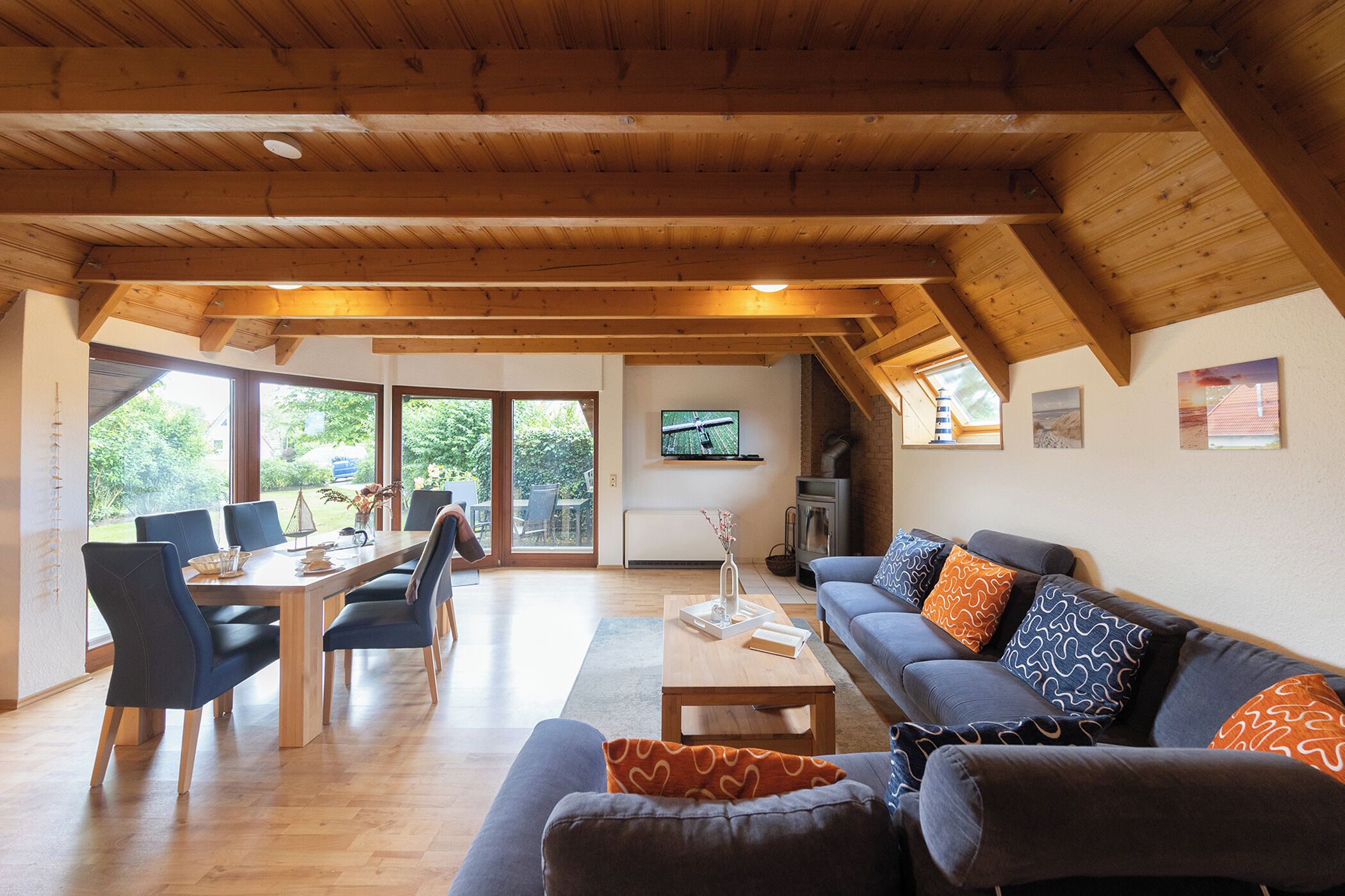 Ein geräumiger Wohnbereich mit Holzbalkendecke, einem Esstisch mit Stühlen, einer Couch mit gemusterten Kissen und einem an der Wand befestigten Fernseher.