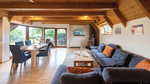 A spacious living area with a wooden ceiling, a dining table with chairs, a sofa with patterned cushions, and a television on the wall. - Vacation home Lichtes Haus am Feld. With two terraces and close to the coast. (Wurster Nordseeküste)