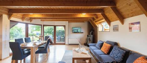 Ein geräumiger Wohnbereich mit Holzbalkendecke, einem Esstisch mit Stühlen, einer Couch mit gemusterten Kissen und einem an der Wand befestigten Fernseher.