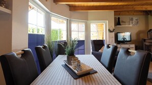 Uma sala de jantar com mesa posta para seis pessoas, cercada por cadeiras, uma televisão e uma lareira.
