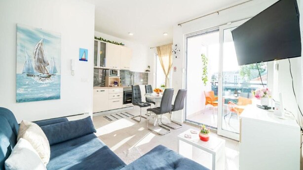Een moderne woonkamer met een bank, een eethoek met stoelen, een keuken op de achtergrond, en een groot raam met uitzicht.