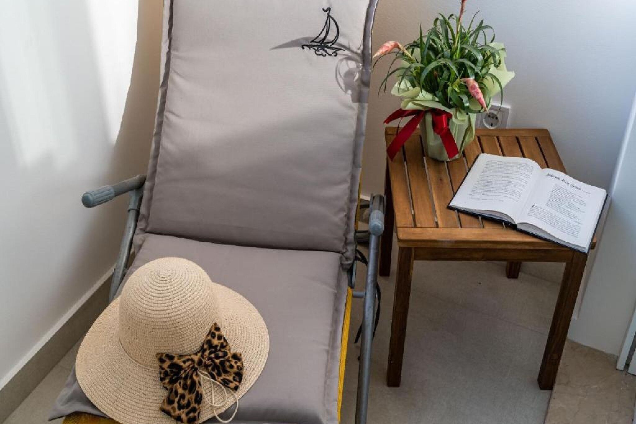 A reclining chair with a cushion, a wooden table with a book, a potted plant, and a straw hat with a bow.