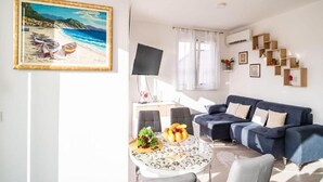Een woonkamer met een blauwe bank, een eettafel met fruit, en een schilderij van een strandgezicht.