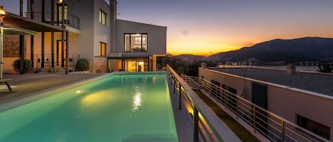 A modern two-story house with a swimming pool, a balcony, and a view of the sunset over the hills.