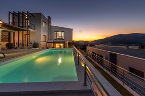 Une maison moderne à deux étages, dotée d’une piscine, d’un balcon et offrant une vue sur le coucher de soleil sur les collines.