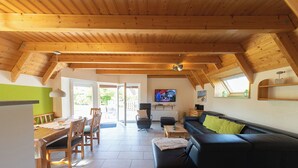 A cozy living room with a wooden ceiling, a black sofa with a green pillow, a dining table with chairs, and a TV on the wall.