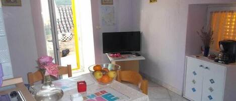 Uma sala de jantar com uma mesa posta para dois, uma televisão em um móvel e um armário com gavetas.