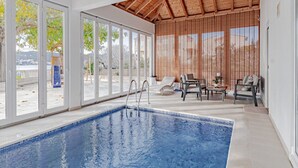 Une piscine intérieure dotée d’un plafond en bois, de grandes fenêtres et d’un espace détente avec des chaises et une table.