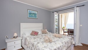 Un dormitorio con cama, mesita de noche, lámpara, un cuadro y vista al mar a través de un ventanal corredero.