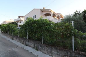 Residential building with a two-story structure and a balcony on the upper floor, surrounded by lush greenery and a low stone wall.