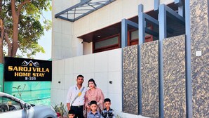 Uma família em frente a um prédio com uma placa indicando "SAROJ VILLA HOME STAY B-225".