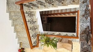Uma escadaria com corrimão de madeira e guarda-corpos de metal, que leva a um cômodo escuro com uma janela.