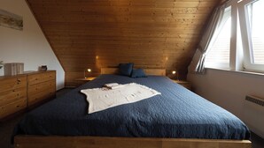 A bedroom with a sloped ceiling, wooden headboard, a large bed with a blue quilt, two bedside tables with lamps, and a window with curtains.