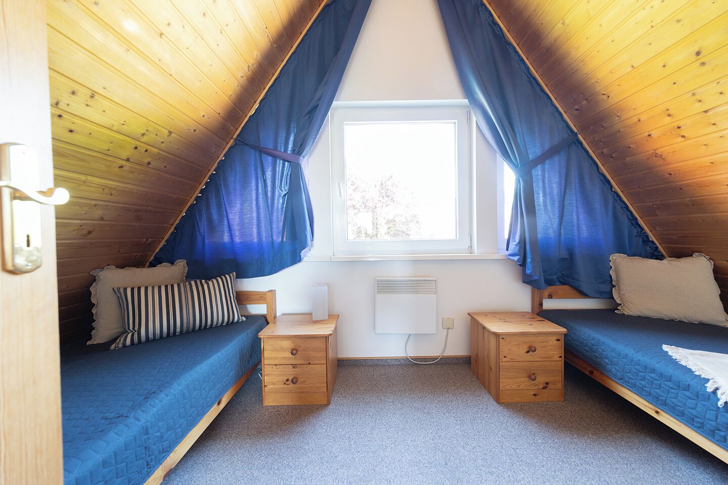 A small, cozy attic bedroom with two single beds, a window with curtains, a heater, and a small wooden nightstand.