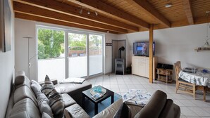 A living room with a wooden ceiling, a sofa, a coffee table, a television, and a dining area with a table and chairs. - Vacation home Ebb & Flow. Bright and tasteful. (Wurster Nordseeküste)