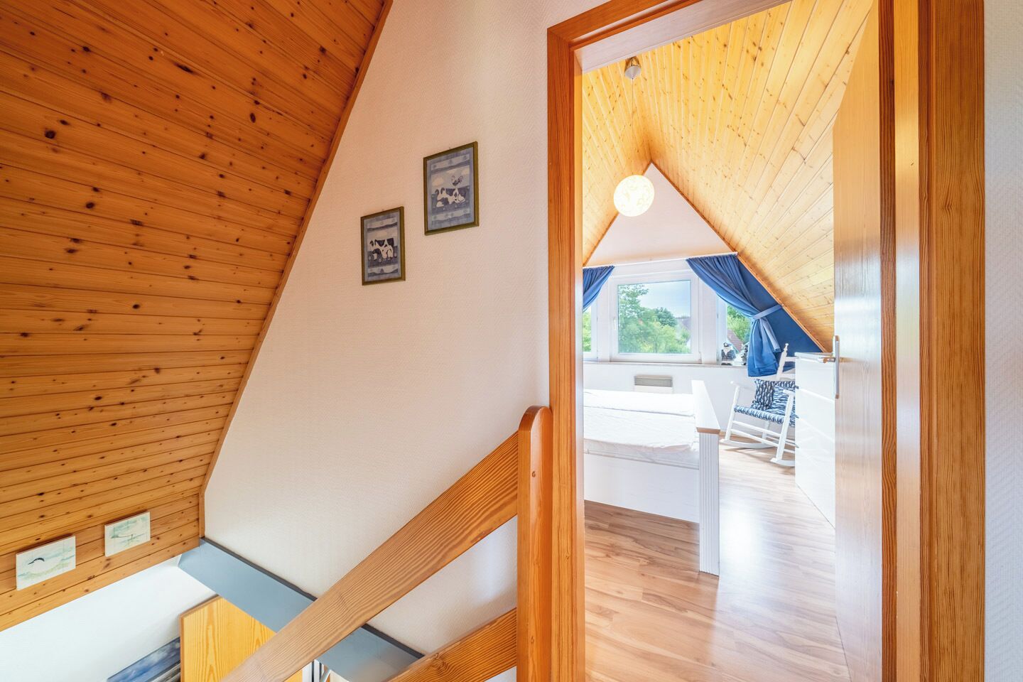 A wooden interior with a sloped ceiling, a staircase, and a view into a room with a bed and a chair.
