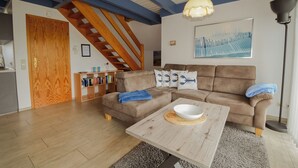 Een woonkamer met een bruine bank, een houten salontafel en een trap met houten balken.