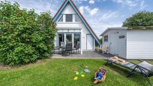 Een huis met schuin dak, een terras met tafel en stoelen, en een tuinhuis in de achtertuin.