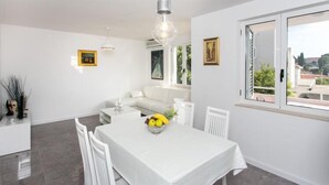 A dining area with a white table, chairs, and a fruit bowl. There is a white sofa and a window with a view of buildings.