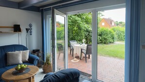 Un salon moderne avec un canapé bleu, une table basse sur laquelle se trouve un saladier de pommes, et une porte-fenêtre coulissante donnant sur une terrasse avec une table et des chaises.