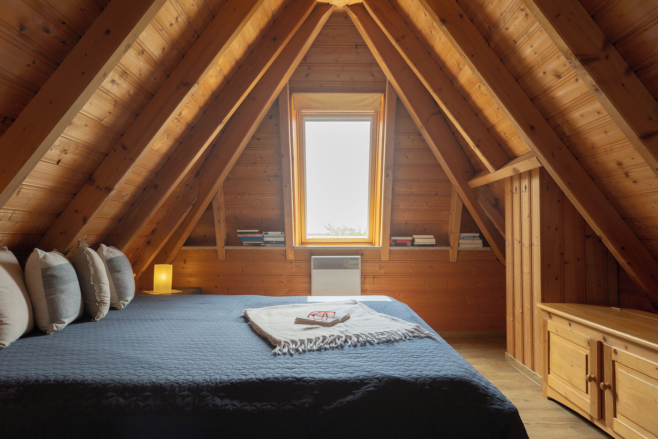 Een gezellig slaapkamer op zolder met een schuine plafond, een groot raam, een bed met kussens, een nachtkastje met een lamp en een houten dressoir.