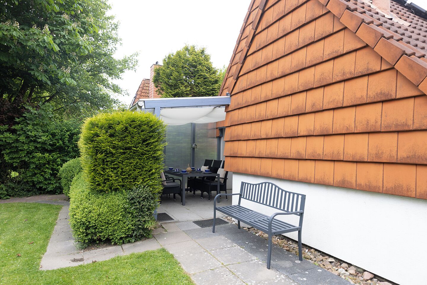 A patio area with a table and chairs, a bench, and a tall, neatly trimmed hedge.