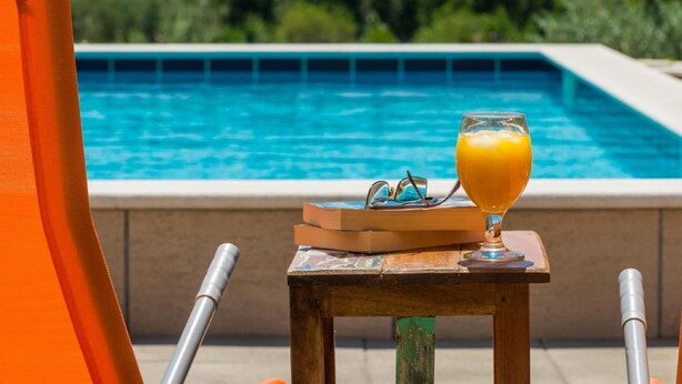 Une scène de bord de piscine avec un verre de jus d’orange, des lunettes de soleil et un livre posé sur une petite table.