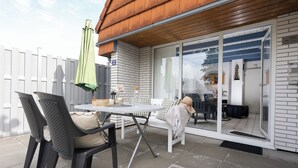 Eine Terrasse im Freien mit Tisch und Stühlen, einem grünen Sonnenschirm und Blick ins Haus durch Glastüren.