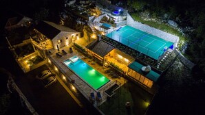 Vista aérea de una propiedad con piscina, cancha de tenis y edificio de piedra con un nivel superior iluminado en azul.