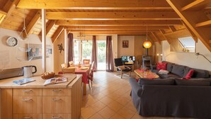 Une pièce chaleureuse et bien éclairée, comprenant un coin repas, une cuisine et un espace salon, avec des poutres en bois et une grande fenêtre.