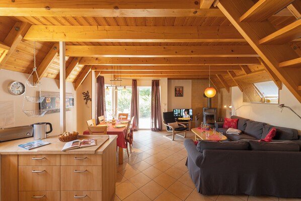 A cozy, well-lit room with a dining area, kitchen, and seating space, featuring wooden beams and a large window.