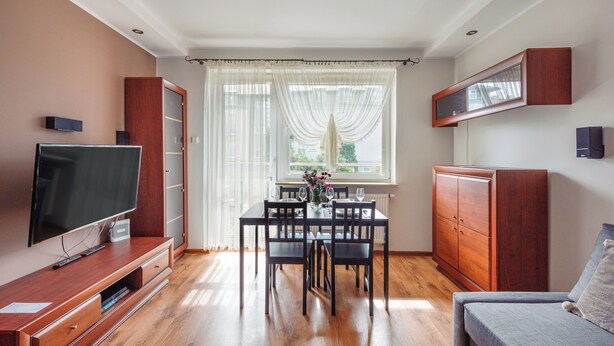 A modern living room with a flat-screen TV, wooden cabinets, a dining table set for four, and a large window with sheer curtains. - Apartament Sun & Snow Siedleckiego 8 (Wladyslawowo)