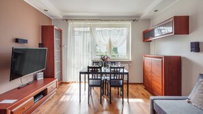 A modern living room with a flat-screen TV, wooden cabinets, a dining table set for four, and a large window with sheer curtains. - Apartament Sun & Snow Siedleckiego 8 (Wladyslawowo)