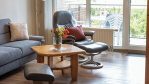 A modern living room with a grey sofa, a wooden coffee table, a leather armchair, and a view of the outdoors through large windows.